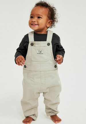 Lachende peuter met krullend haar, gekleed in beige tuinbroek en een zwart shirt met lange mouwen, staat blootsvoets tegen een effen achtergrond.