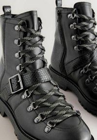 Fekete bokacsizmák texturált műbőr kígyóbőr részlettel, fényes fém fűzőlyukakkal és keresztbefonott fűzőkkel, az egyik oldalon cipzárral.