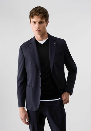 Giovane uomo con capelli ricci che indossa un blazer blu scuro, un maglione nero a scollo a V e una canottiera bianca su uno sfondo chiaro e uniforme.