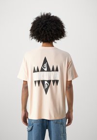 Short-sleeve peach t-shirt featuring a large black and white geometric logo with textured accents on the back. Casual fit, cotton material.