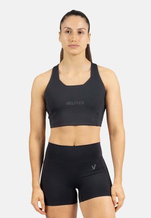 Mujer con cabello oscuro recogido en una coleta, vistiendo un sujetador deportivo negro y pantalones cortos deportivos negros de talle alto contra un fondo blanco liso.