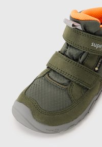 Chaussure verte imperméable avec des sangles Velcro, un tissu texturé et un col rembourré orange. Dotée d'une semelle en caoutchouc avec la marque "superfit".