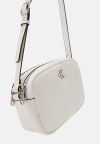 Bílá kožená kabelka přes rameno s texturovaným povrchem, obdélníkovým tvarem, stříbrnými detaily, zipy a decentním logem na přední straně.