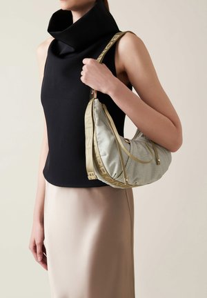 Light green textured shoulder bag with gold hardware and a logo strap, held by a person wearing a black sleeveless top and beige skirt.