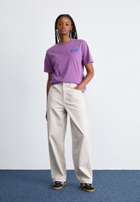 T-shirt en coton violet avec logo bleu, associé à un pantalon large beige clair et des chaussures noires avec semelles jaunes, sur fond neutre.