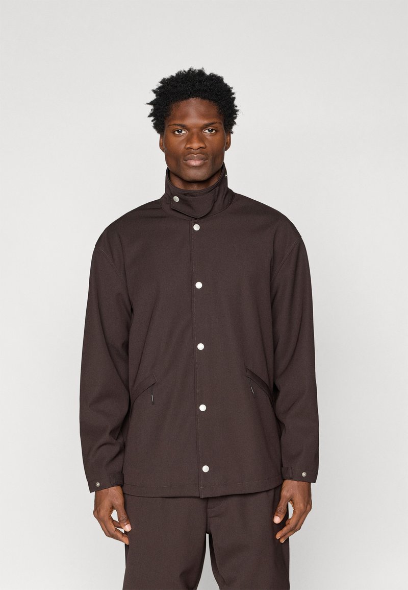 Brown collarless jacket with snap buttons, side pockets, and a relaxed fit. Made from textured fabric, offering a sleek, minimalistic design.