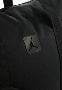Bolsa de deporte negra con material de nailon texturizado, que presenta un parche rectangular negro con el logo de una figura saltando. Con los detalles de correas resistentes.