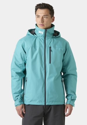 Hombre vestido con una chaqueta impermeable color verde azulado con capucha y bolsillos con cremallera, de pie frente a un fondo claro y llano.