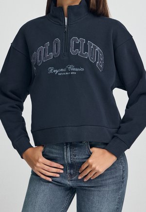 Kobieta ubrana w czarną bluzę z suwakiem do ćwiartki z napisem „POLO CLUB Beyond Classics Established 2012” z przodu i niebieskie dżinsy, ręce spoczywają blisko kieszeni.