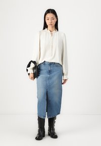 Bílé pruhované blouse s volánkovým límcem, v kombinaci s modrou džínovou sukní s předním rozparkem. Modelka drží černou tašku a nosí vysoké boty po kolena.
