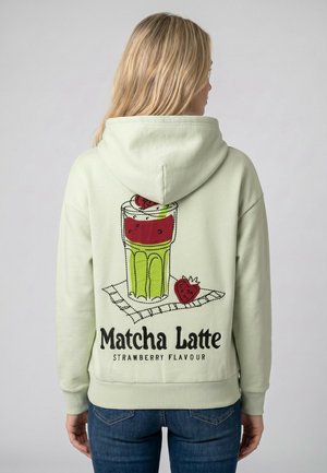 Blonde Person trägt einen hellgrünen Hoodie mit bunter Matcha-Latte- und Erdbeer-Grafik und dem Text "Matcha Latte Strawberry Flavour" auf dem Rücken.
