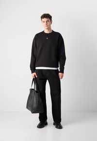 Sudadera negra con un pequeño logo, combinada con pantalones negros. El modelo lleva un bolso tote de cuero negro con un diseño texturizado.