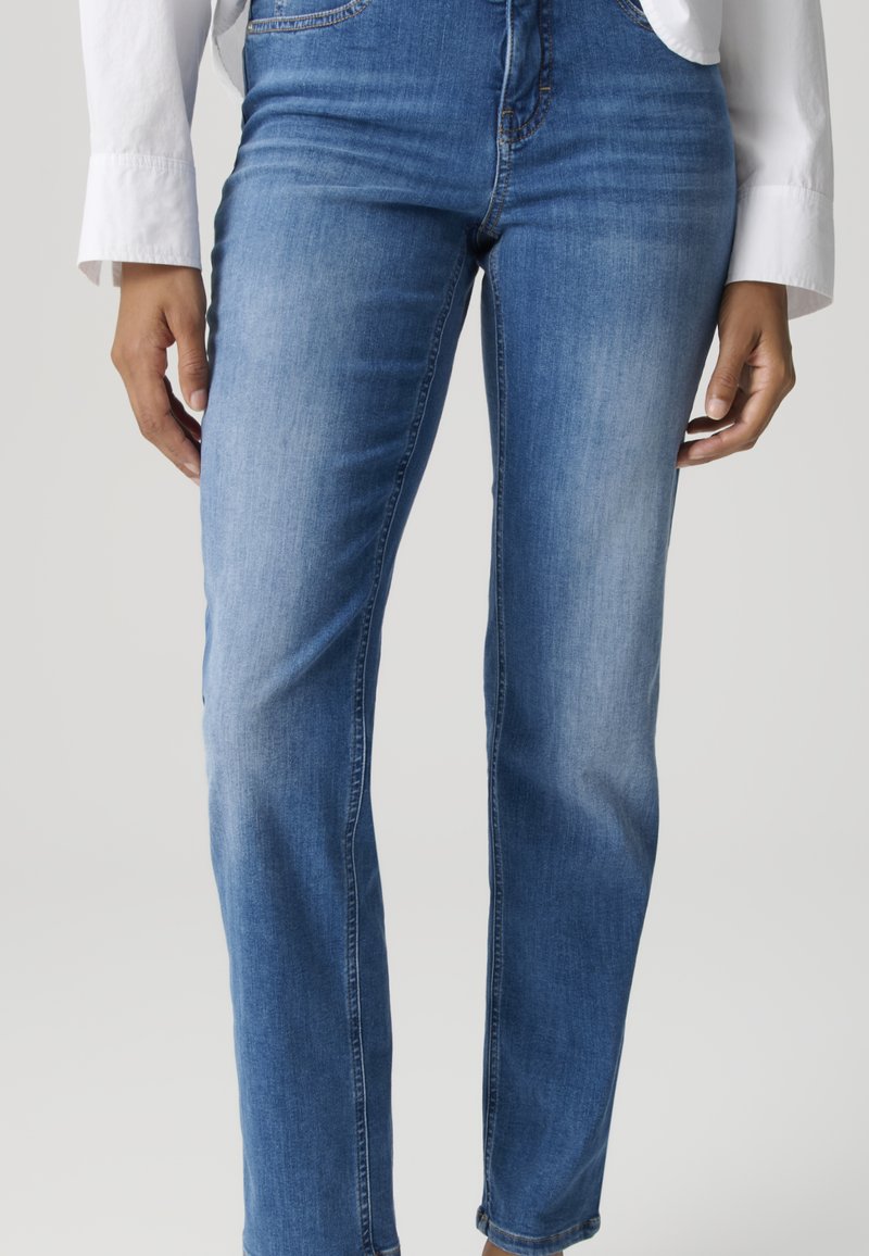 Persona che indossa jeans blu medio a gamba dritta e una camicia bianca a maniche lunghe, in piedi con le mani rilassate ai lati.