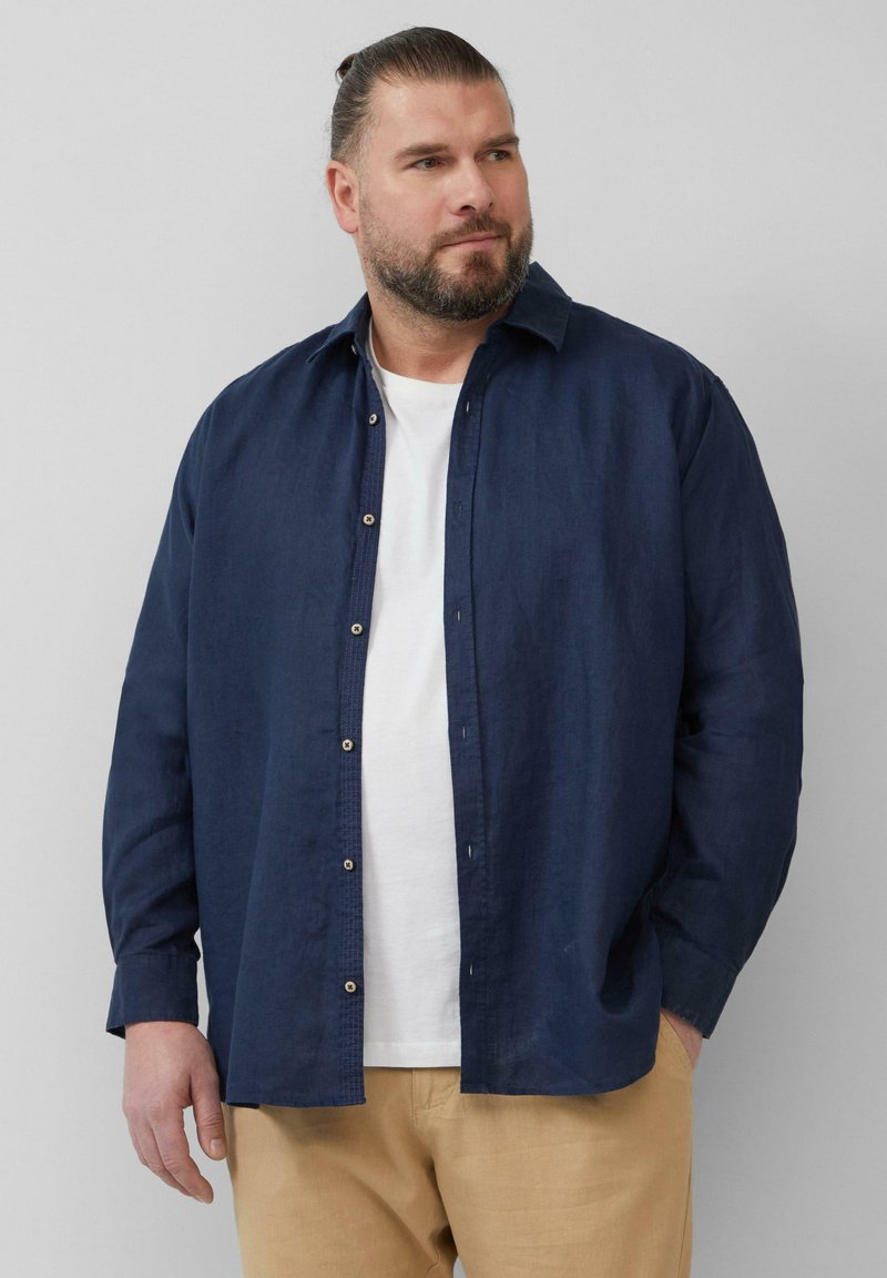 Donkerblauw button-up overhemd van lichtgewicht stof, met een klassieke kraag en lange mouwen, gedragen met een wit T-shirt eronder.