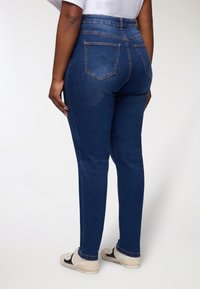 Jeans in denim blu scuro con vita alta, due tasche posteriori e vestibilità slim. Indossati con sneakers di colore chiaro.