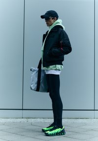 Zwarte bomberjack met textuurstof, muntkleurige hoodie eronder, zwarte leggings en multicolour Nike-schoenen. Grijze schoudertas met zwarte banden.