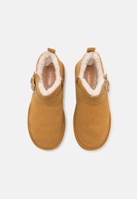 Botas de tobillo de ante marrón con un suave forro de piel sintética blanca, punta redondeada y un cinturón ajustable en el costado. Suela de goma plana.