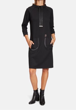 Mujer con vestido negro con capucha hasta la rodilla, bolsillos delanteros y texto "COZY", combinado con botines negros.
