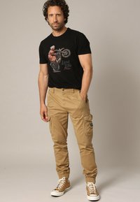 Man met krullend haar in een zwart graphic t-shirt met een motorrijder, beige cargobroek en beige hoge sneakers, staand met één hand in zijn zak.
