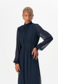 Μακριά φόρεμα ναυτικού μπλε σε ελαφρύ ύφασμα, με ψηλό φουσκωτό λαιμό, μακριά διάφανη μανίκια και ζώνη που δένει στη μέση.