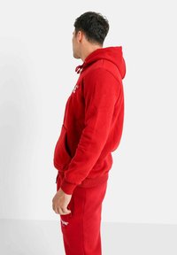Sudadera con capucha roja hecha de un tejido suave, con un bolsillo tipo canguro y puños acanalados. Combinada con pantalones rojos a juego, vista de lado.