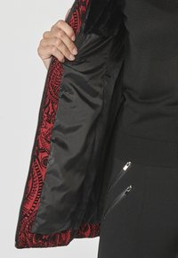 Crna jakna s podstavom od crvenog paisley uzorka, koja ima zatvaranje na patentni zatvarač i elegantnu crnu unutrašnjost. Oslikavaju je vidljivi džepovi sa patentnim zatvaračima.