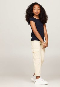 Tumši zils kokvilnas īsroku tops, bēša krāsas kaujas stila bikses ar sānu kabatām un balti sporta apavi. Meitene stāv ar vaļīgi noliktām rokām gar sāniem.