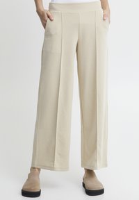 Beige vide bukser med en glat tekstur, der har frontfolder og sidelommer. Kombinert med lysebrune slip-on sko.