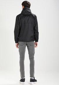 Zwarte waterdichte jas met capuchon, elastische manchetten en een losse pasvorm. Gezien van achter, gedragen over grijze jeans en zwarte sneakers.