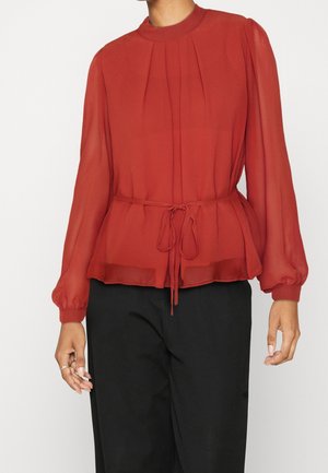 Femme portant un chemisier rouge à manches longues avec des poignets froncés et une ceinture à la taille, associé à un pantalon noir ample.