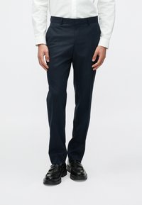 Tengerészkék szabott nadrág sima textúrával, rögzített derékpánttal és oldalzsebekkel, fekete bőrcipővel párosítva.