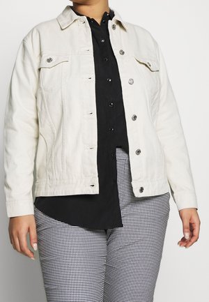 Biała kurtka dżinsowa z przodem na guziki, dwoma kieszeniami na piersi i srebrnymi dodatkami, nałożona na czarną koszulę i spodnie w wzór pepitki.