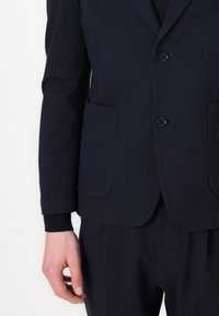Navy blazer in smooth fabric, featuring a two-button closure, notched lapels, and two front pockets. Cuffs with black ribbed texture.
