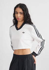 Ženska z dolgimi kodrastimi lasmi, oblečena v bel kratki Adidas top z črnimi črtami in črne hlače z visokim pasom, stoji pred enostavnim ozadjem.