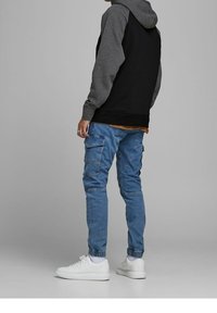 Persona che indossa una felpa con cappuccio nera e grigia, jeans cargo blu e sneakers bianche, in piedi davanti a uno sfondo grigio uniforme.