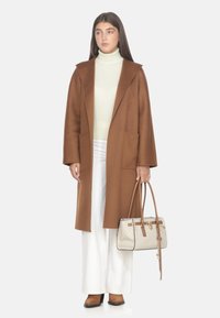 Donna che indossa un lungo cappotto marrone, un maglione a collo alto color crema, pantaloni bianchi, stivali marroni, mentre tiene una borsa beige e marrone con manici lunghi.
