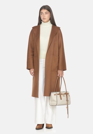 Donna che indossa un lungo cappotto marrone, un maglione a collo alto color crema, pantaloni bianchi, stivali marroni, mentre tiene una borsa beige e marrone con manici lunghi.