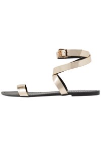 Guldmetalliska platta sandaler med svart sula. Har två tunna remmar och ett spänne vid ankeln. Enkel design, slät textur, öppen tå.