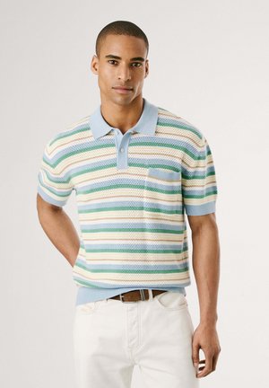Polo de rayas en azul, verde y beige, con un tejido texturizado, cuello, tres botones y un bolsillo en el pecho. Se lleva con pantalones blancos.