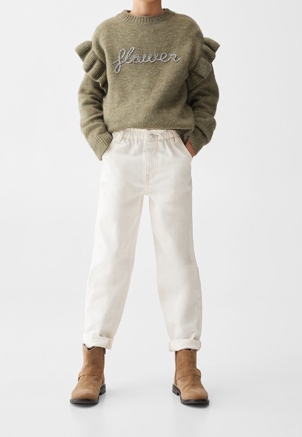 Enfant portant un pull vert olive à manches volantées avec le texte "flower", pantalon blanc retroussé et bottines couleur sable avec les mains dans les poches.