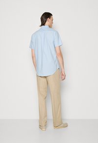 Camisa de manga curta azul clara com um pregueado nas costas, combinada com calças bege e calçado bege, em fundo branco liso.