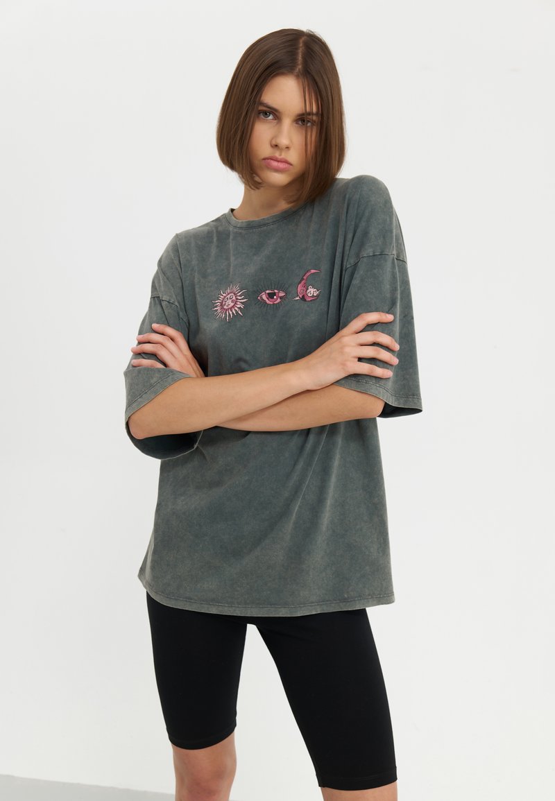T-shirt oversize gris à manches courtes, avec des détails graphiques de soleil et de lune roses. Associé à des shorts de vélo noirs. Texture lisse.