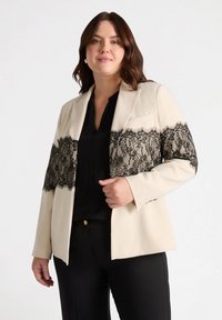 Blazer crema con detalles de encaje negro en el pecho, con un corte entallado, solapas con muesca y mangas largas.