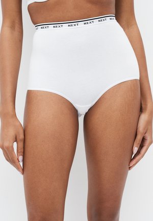 Frau trägt weiße Baumwoll-Slips mit hohem Bund und elastischem Taillenbund, der mit der Aufschrift "NEXT" versehen ist, vor einfarbigem Hintergrund.