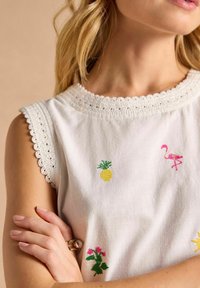 Helle ärmellose Bluse mit einem gehäkelten Ausschnitt, verziert mit bunten Stickereien eines Ananas, eines Flamingos und Blumen in einem verspielten Design.