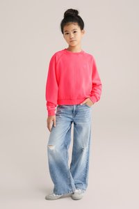 Ryškiai rožinė džemperis su raukiniais ant rankovių ir apačios, derinamas su šviesiai mėlynomis plačiomis kelnėmis, turinčiomis nusidėvėjimo elementų. Su stilizuotais pilkais batais.