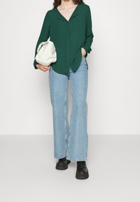 Blusa verde a maniche lunghe con scollo a V e spacchi laterali, abbinata a jeans svasati blu chiaro e stivali platform neri, mentre tiene una borsa bianca.