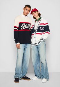 Navy and cream knit sweater with red stripes and logo; white, red, and navy cardigan; blue wide-leg jeans; dark and white sneakers.