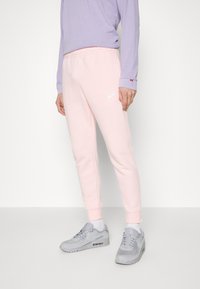 Světle růžové tepláky s tapered designem, elastickým pasem a bílým logem Nike. V kombinaci se šedými teniskami a fialovým tričkem s dlouhým rukávem.