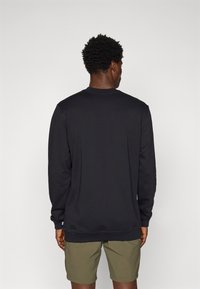 Sudadera negra de manga larga con puños acanalados, con un cuello redondo y un pequeño logo en el dobladillo. Lleva puesta con pantalones cortos verdes.